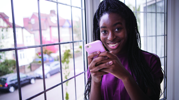Young woman stands holding her phone and smiles towards the camera